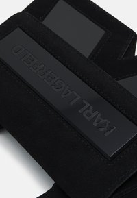 Zwarte suède product met een glanzend, gegraveerd zwart hardwaredetail. Heeft een rechthoekige vorm en stiksel langs de randen.