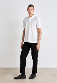 Mann som står i hvit kortermet polo skjorte, svarte bukser, svarte sokker og svarte slip-on sko mot en ensfarget vegg og gulv bakgrunn.