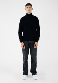 Czarny sweter z golfem wykonany z miękkiego materiału, w połączeniu z ciemnoszarymi dżinsami i białymi sneakersami. Model stoi na jednolitym tle.