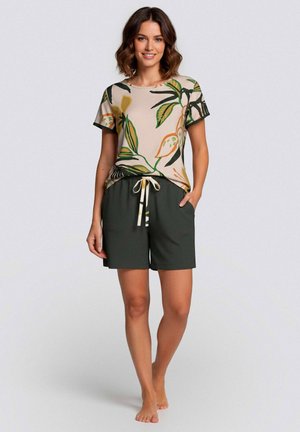 Vrouw staat blootsvoets en draagt een beige top met groene en oranje bladpatronen en donkergroene korte broek met een trekkoord in de taille.