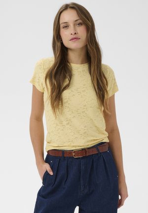 Kvinde med langt brunt hår iført en gul blonder kortærmet top, blå bukser med høj talje og et brunt bælte, stående med den ene hånd i lommen.