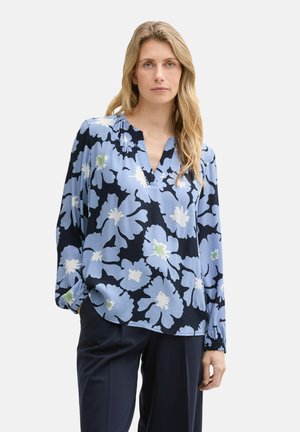 Langsleeve blouse in blauw met een groot bloemenpatroon in wit en groen, met een V-hals en een geplooide halslijn, gecombineerd met donkere broeken.