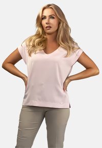 Jasnoróżowa bluzka z krótkim rękawem, dekoltem w kształcie litery V, gładką fakturą, stonowanym designem i małym, srebrnym akcentem marki przy dolnym szwie.