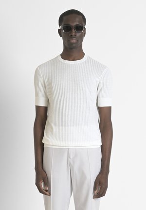 Maglione bianco a coste con maniche corte e scollatura rotonda, abbinato a pantaloni grigi chiari. Il modello indossa occhiali da sole neri. Maglione in maglia morbida e testurizzata.
