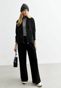 Zwarte zip-up hoodie met trekkoorden, grijze coltrui en losse zwarte broek. Grijze gebreide beanie en kleine zwarte handtas. Witte sneakers.