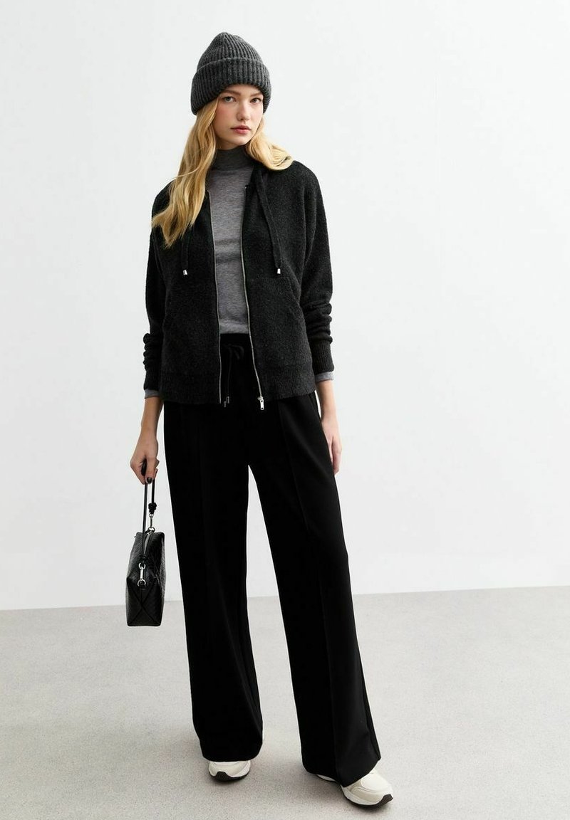 Felpa nera con zip e cappuccio regolabile, dolcevita grigia e pantaloni neri larghi. Berretto grigio lavorato a maglia e piccola borsa nera. Sneakers bianche.