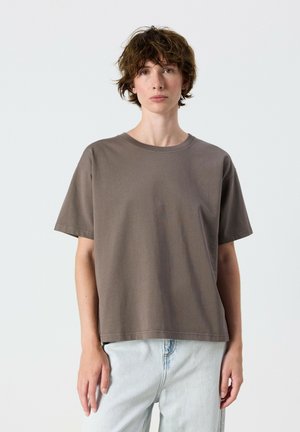 Persona con capelli corti, castani e disordinati, che indossa una maglietta marrone larga e jeans azzurri, in piedi davanti a uno sfondo semplice.