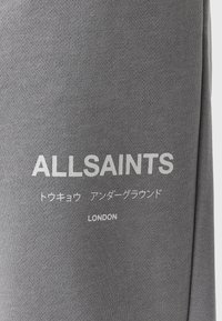 Țesătură gri cu text alb care spune ALLSAINTS, caractere japoneze și LONDON imprimat pe ea.