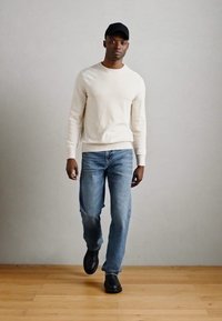 Bílé pletené svetr, modré džíny, černá kšiltovka a černé boty. Jednoduchý, neformální outfit s volným střihem a hladkou texturou.