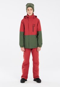 Rote und grüne wasserdichte Jacke mit Kapuze, Reißverschlusstasche und schwarzen Akzenten. Kombiniert mit roten Hosen und schwarzen wasserdichten Stiefeln.