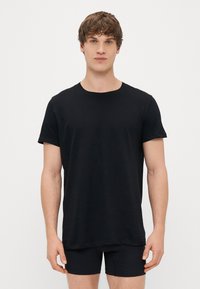 Juodas medvilninis marškinėliai su apvaliu kaklu ir trumpomis rankovėmis, turintis tiesų apatinį kraštą ir laisvą siluetą, dėvimi virš juodų apatinukų.