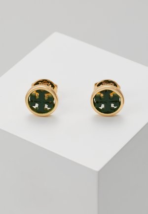 Złote kolczyki na sztyft z zielonym emaliowanym inkrustacją z wyciętym geometrycznym wzorem, prezentowane na białym pedestalzie.
