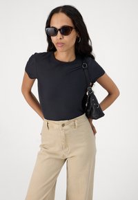 Schwarzes kurzärmeliges Oberteil kombiniert mit hochgeschnittenen beigefarbenen Hose; das Model hält eine kleine schwarze Tasche und trägt eine dunkle Sonnenbrille.