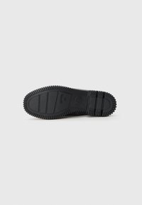Svart gummisåle med teksturert grep, med rygger og hevede seksjoner for bedre traction. Solid og fleksibel design.