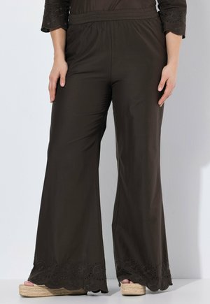 Frau trägt dunkelbraune, weite Hose mit bestickten, gewellten Säumen und beigefarbenen Keilsandalen, steht vor einer schlichten hellen Wand.