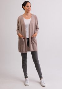 Beige cardigan met lange mouwen, open voorkant en twee voorzakken. Gecombineerd met grijze skinny jeans en witte sneakers.
