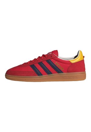 Zapatilla Adidas Spezial roja con rayas negras, suela de goma y lengüeta amarilla en el talón, vista de perfil sobre un fondo blanco.