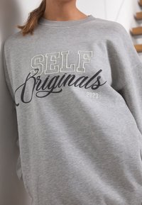 Felpa grigia con la scritta "SELF Originals 2023" stampata in bianco e nero, con scollo rotondo e vestibilità comoda. Texture morbida visibile.