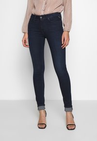 Skinny dark blue jeans with a classic five-pocket design. The hem is rolled up, showcasing black strappy sandals. The fabric has a smooth texture.