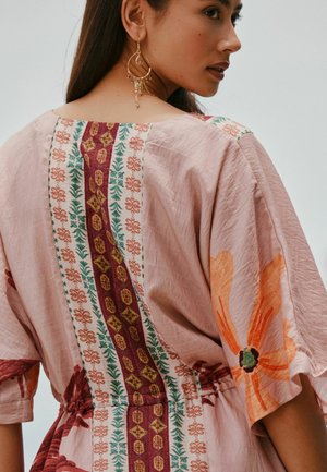 Frau in einem hellrosa Kleid mit floralen und geometrischen Mustern, von hinten gezeigt und über die Schulter blickend, trägt hängende Ohrringe.