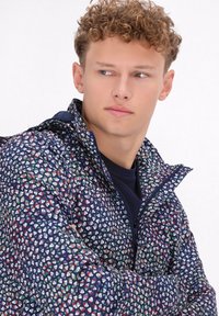 Jachetă marine cu un model geometric multicolor, închidere cu fermoar, guler înalt și o ajustare adaptabilă, realizată dintr-un material ușor și texturat.