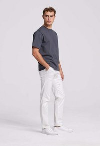 Grijze katoenen T-shirt met korte mouwen, gecombineerd met witte jeans en witte sneakers. Model poseert met handen in de zakken tegen een effen achtergrond.