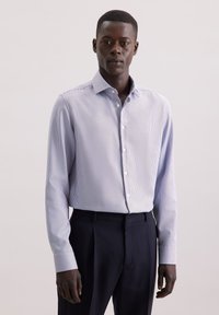 Camicetta da uomo a maniche lunghe di colore azzurro chiaro con un motivo a piccoli quadretti, caratterizzata da un colletto button-down e tessuto morbido, abbinata a pantaloni scuri.