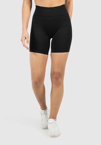 Vrouwelijk model in zwarte shorts en witte sneakers, aan het lopen voor een effen achtergrond. Focus op de details van de benen en de outfit.