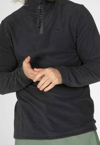 Pull noir en polaire avec un col zippé, arborant un logo sur la poitrine et un tissu texturé. Manches longues avec une coupe décontractée.