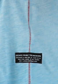 Hellblauer Stoff mit einer markanten roten Naht in der Mitte. Ein schwarzes Etikett mit weißem Text ist sichtbar und enthält Markeninformationen.