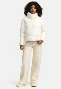 Doudoune blanche avec un col haut, un design à coutures horizontales et des fermetures à boutons. Associée à un pantalon large crème et des baskets beiges.