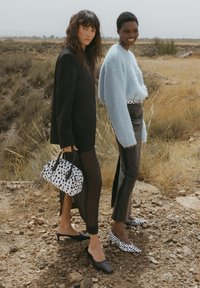 Dwa modele stoją w suchej okolicy. Jeden z nich nosi czarną marynarkę i czarne spodnie, a drugi ma na sobie jasnoniebieski sweter i skórzane spodnie w kropki. Trzymają małą torebkę w czarno-białe kropki. Obydwoje mają na sobie stylowe buty: jeden w czarnym kolorze, a drugi w białym z wzorem w kropki.