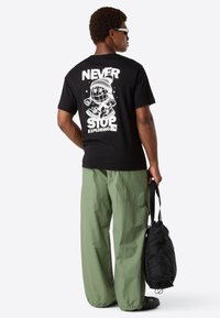 Man wearing sunglasses, black graphic tee with "Never Stop Exploring," loose green pants, holding a black bag against a white background.