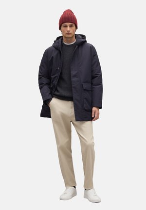 Marinefarbener, gepolsterter Mantel mit Kapuze, grauer Pullover, cremefarbene Hose und weiße Sneaker. Dunkelrote Strickmütze. Hat einen Reißverschluss und einen Knopfverschluss sowie Seitentaschen.