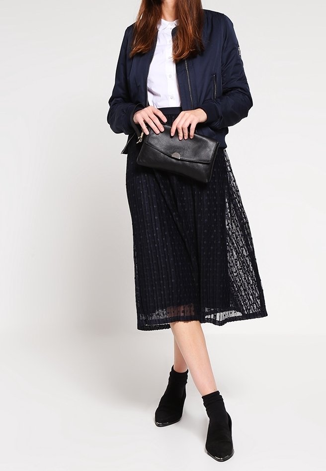Femme portant une veste bomber bleu marine, une blouse blanche, une jupe midi bleu marine transparente, des bottines noires, tenant une pochette noire.