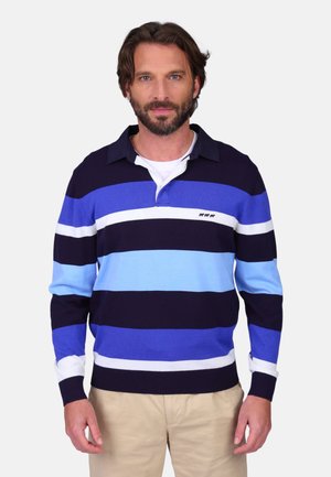 Homme barbu portant un maillot de rugby à rayures bleues, noires et blanches, et un pantalon beige, debout devant un fond blanc uni.