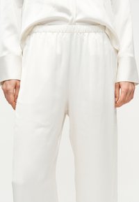 Oseba v širokih belih hlačah z elastičnim pasom in ujemajoči se beli majici z dolgimi rokavi, roke sproščene ob telesu.