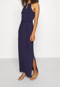 Mörkblå maxiklänning med halterneck och nyckelhålsdetalj, med en åtsittande midja och sidofickor, tillverkad av ett mjukt tyg.