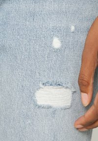 Ljusblå slitna denim med fransade kanter och flera revor, med en närbild som betonar textur och detaljer.