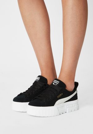 Musta mokkanahkainen korollinen tennari, jossa on valkoinen korosteraita ja paksu valkoinen kumipohja, jossa on kuvioidut urat, ja siihen sopivat mustat nauhat.