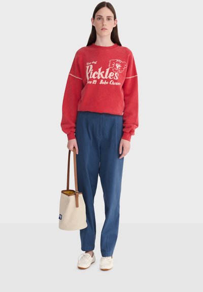 Kobieta w czerwonym swetrze z białym napisem i grafiką, niebieskich spodniach, białych butach, trzymająca beżową i brązową torebkę na jednolitym tle.