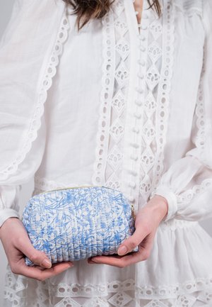 Pochette patchwork bleu à motif floral avec fermeture éclair dorée, tenue dans les mains. Le matériau semble léger avec une texture douce. Forme arrondie.
