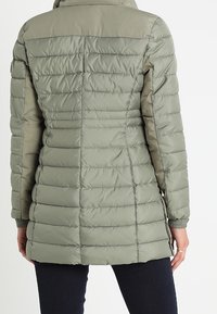 Chaqueta acolchada de color verde oliva que presenta un patrón de acolchado horizontal, cuello alto y textura suave. Incluye puños elásticos y un diseño con la parte trasera más larga.