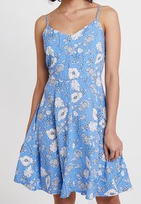 Kvinna i en ärmlös blå klänning med tunna band och vitt blommönster, stående framför en enfärgad ljus bakgrund.