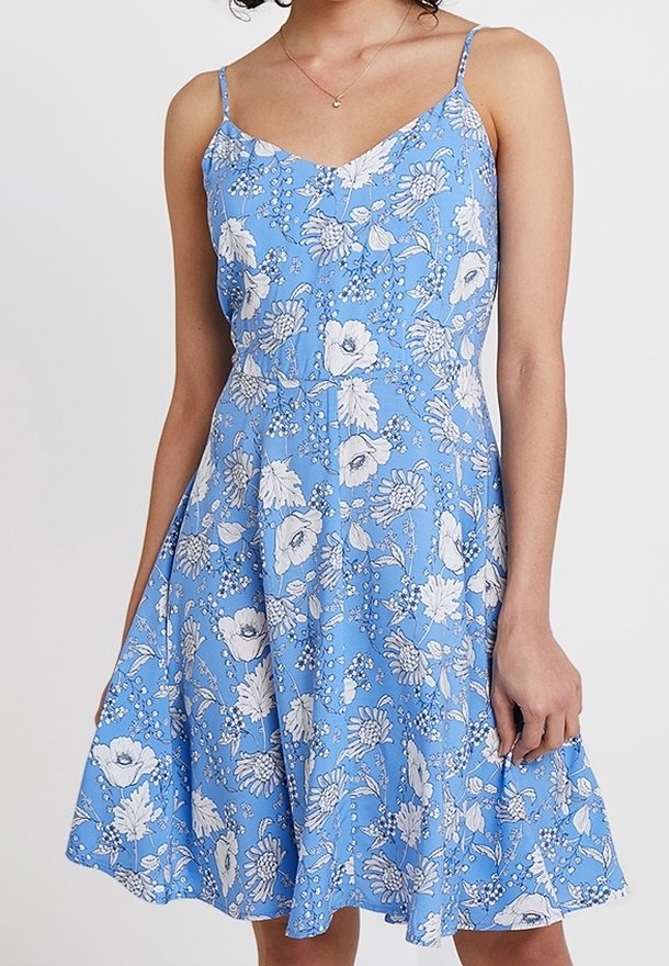 Kvinna i en ärmlös blå klänning med tunna band och vitt blommönster, stående framför en enfärgad ljus bakgrund.