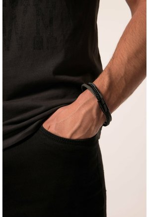 Black T-shirt with subtle graphic texture, paired with dark pants. A braided black bracelet with a metallic clasp is worn on the wrist.