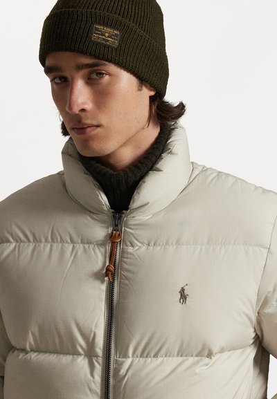 Casaco puffer bege com gola alta, fecho de zíper e cordão castanho. Gorro de malha canelada verde-escuro com um rótulo na frente.