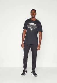 Μαύρο μπλουζάκι με το λογότυπο "ADIDAS SPORTSWEAR" σε λευκό και γκρι, συνδυασμένο με μαύρο παντελόνι φόρμας και μαύρα αθλητικά παπούτσια με λευκές λεπτομέρειες.