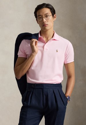 Férfi, aki kerek szemüveget visel és rózsaszín galléros pólót, tengerészkék zakót tart a vállán, egyik keze tengerészkék redős nadrág zsebében.