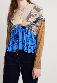 Multifunkční blůzka s výstřihem do V, zdobená květinovými vzory v modré, béžové a hnědé barvě. Dlouhé rukávy a rozšířený střih s nařaseným materiálem.